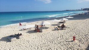 Une plage avec des parasols blancs et des chaises longues, une tour de surveillance pour les sauveteurs, et une vue dégagée sur l’océan.