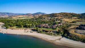 Vista aérea de un complejo turístico costero con villas frente a la playa, una playa de arena y colinas circundantes.