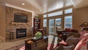Un salón con chimenea de piedra, televisor de pantalla plana, estantería, mesa de comedor con sillas, sofá y vistas a través de amplias ventanas.