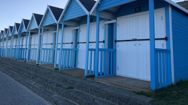 Eine Reihe von blau-weißen Strandhütten, nummeriert von 50 bis 57, mit Holzstufen, die zu jedem Häuschen führen.