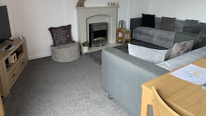 Ein Wohnzimmer mit Kamin, grauem Sofa und einem Fernseher auf einem Holzgestell.
