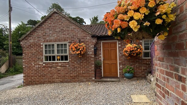 A small brick house with a tiled roof, a front door, and two windows. There are hanging flower baskets on the wall.