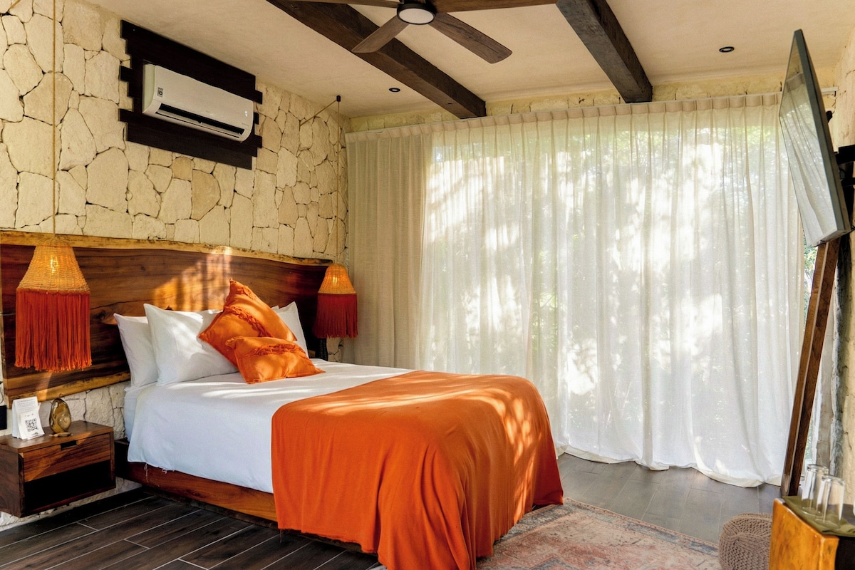 A bedroom with a stone wall, a ceiling fan, an air conditioning unit, a bed with white bedding and orange pillows, a nightstand with a lamp, and a window with sheer curtains.