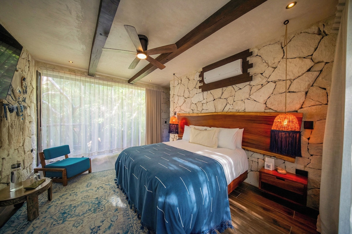 A bedroom with a large bed, a ceiling fan, a stone wall, and a nightstand with a lamp.