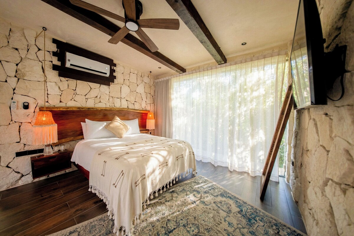 A bedroom with a stone wall, a wooden headboard, a ceiling fan, a bed with white bedding, two bedside lamps, and a large window with sheer curtains.