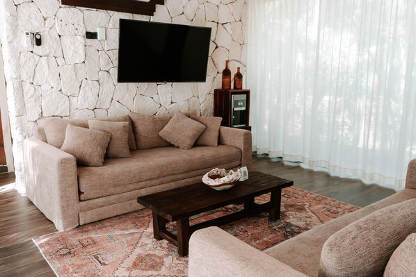 A living room with a stone wall, a flat-screen TV, a sofa, a coffee table, and a rug. - Kaaba Luum Jungle Villas (Tulum)