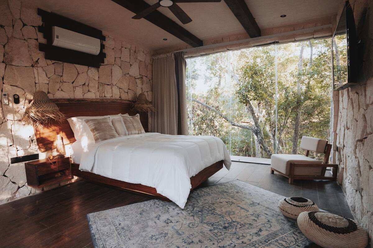 Un dormitorio con pared de piedra, una cama, una mesita de noche, una lámpara, una silla y vistas a árboles a través de un ventanal grande.