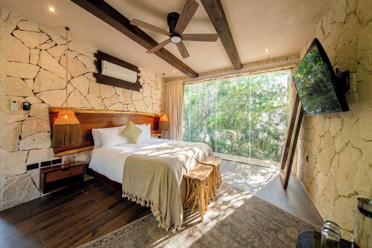 A bedroom with a stone wall, wooden ceiling beams, a bed with a headboard, bedside tables, a nightstand, a large window, and a ceiling fan.