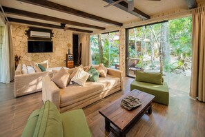 A modern living room with a stone wall, a sofa, a coffee table, and a view of a tropical garden. - Kaaba Luum Jungle Villas (Tulum)