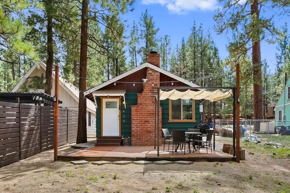 A small, single-story cabin with a brick exterior and a green door, surrounded by a wooden fence and trees. - Moose's Cabin near Big Bear Village and Slopes! (Big Bear)