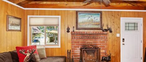 A cozy living room with a fireplace, wooden walls, a ceiling fan, and a couch with decorative pillows.