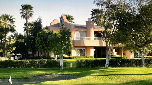 Un campo de golf con un mástil de bandera, césped impecable y un edificio con balcón al fondo.
