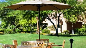 Un patio al aire libre con mesas y sillas de madera, cubierto por grandes sombrillas, con abundante vegetación y un edificio de fondo.