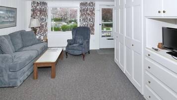 A modern living room with a grey sofa, a round armchair, and a wooden coffee table. There is a TV on a white cabinet, and a window with curtains.