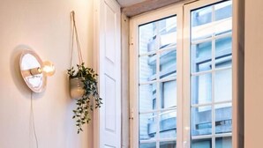 A wicker chair with a yellow cushion and a white pillow, a hanging plant, and a window with blinds. - Lemago Poveiros Apartments (Porto)