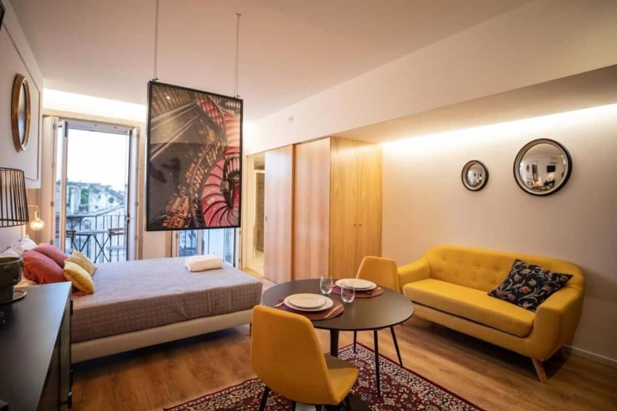 A modern room with a bed, a yellow sofa, a dining table with chairs, and a large abstract painting.