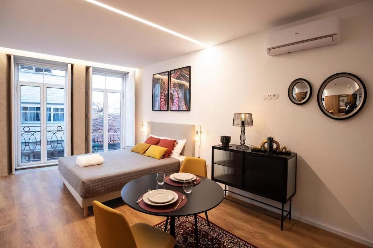 A modern hotel room with a bed, a dining table with chairs, a TV, and artwork on the walls.