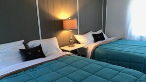 Quarto de hotel com duas camas, cada uma com roupa de cama verde-água e almofadas brancas. Há uma mesa de cabeceira com uma luminária entre as camas.