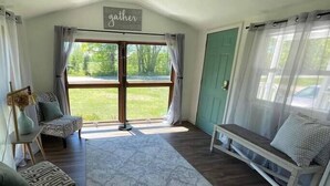 Um quarto espaçoso com uma janela grande, um banco, um tapete estampado e uma placa na parede com a palavra 'gather'.
