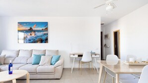 A modern living room with a sofa, a dining table, and a painting of a coastal scene. - Home2Book Comfy Seaside Apartment Cristimar, Pool (Los Cristianos)