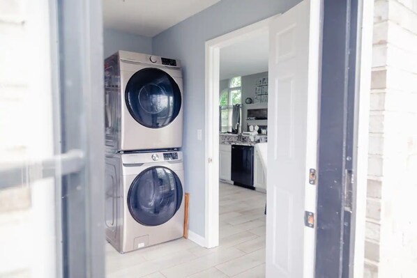 A laundry room with stacked front-loading washers and dryers, a kitchen in the background, and a door leading to another room.