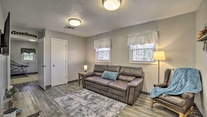 A living room with a brown leather sofa, a blue armchair, a wooden coffee table, and a patterned rug. There are two windows with white curtains, a lamp, and a door labeled "CAMERON". - Near Mines & Meadows: Spacious Home w/ Game Room (Beaver Falls)