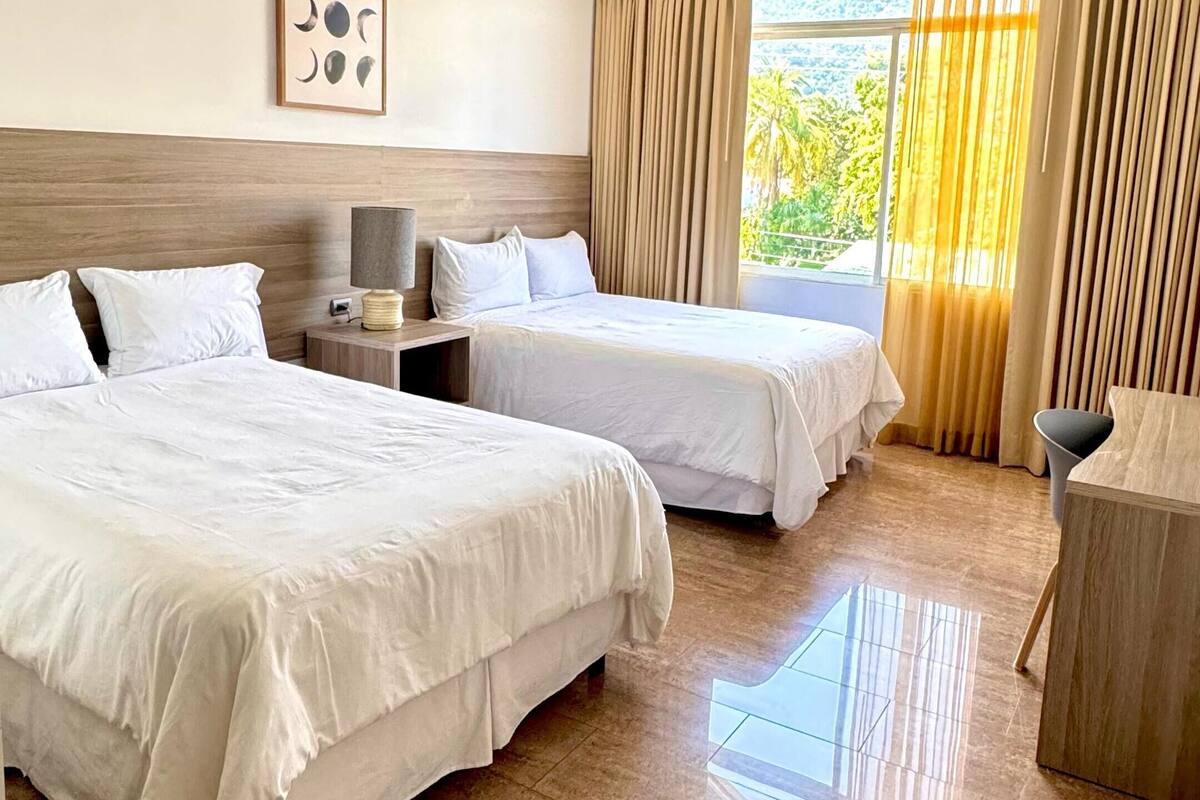 A hotel room with two beds, a nightstand, a lamp, a ceiling-mounted security camera, and a view of greenery through the window.