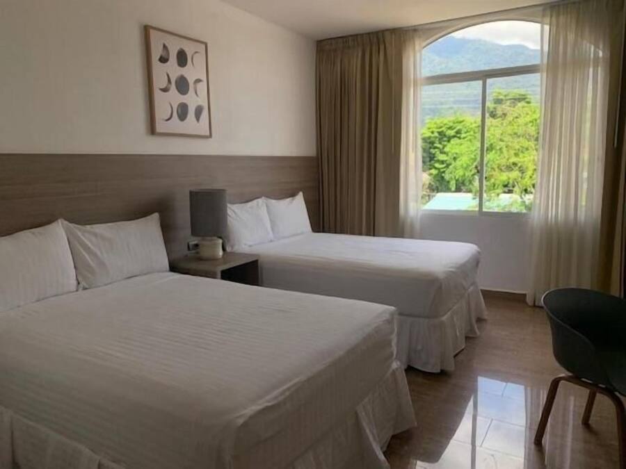 A hotel room with two beds, a nightstand with a lamp, a chair, and a window with curtains.