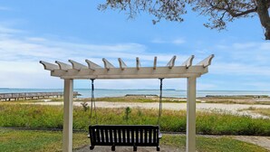 A white pergola with a black bench underneath, overlooking a beach with a wooden boardwalk and the ocean in the background.