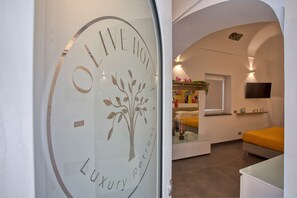 A modern interior with a glass door featuring a logo and the words 'OLIVE HOTEL' and 'Luxury Retreat.' Inside, there is a bed with a yellow headboard, a flat-screen TV mounted on the wall, and a window with a view. - Olive House, a luxurious retreat (furore)