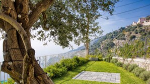 Un giardino curato con un grande albero, una zona pavimentata e vista su una località costiera.