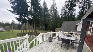 A wooden deck with outdoor furniture, surrounded by a white railing, overlooking a grassy yard with trees. - Charming Gig Harbor Getaway (Gig Harbor)