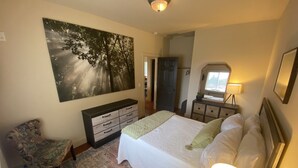 A bedroom with a bed, a nightstand with a lamp, a dresser, a chair, a large painting, and a door leading to another room. - Charming Gig Harbor Getaway (Gig Harbor)