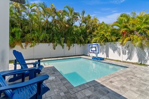 Una piscina con sillas Adirondack azules, un aro de baloncesto y una pared blanca con palmeras.
