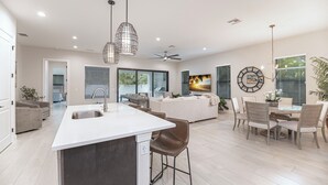 A modern kitchen with a central island, dining area with chairs, and a living room with a sofa and TV. - The Blue Heron | Heated Pool and Outdoor Grill | Near Beach (Naples)