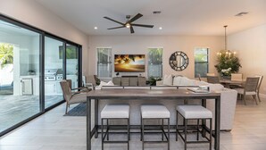 A modern kitchen with a large island, a dining area with a chandelier, and a living room with a large painting. - The Blue Heron | Heated Pool and Outdoor Grill | Near Beach (Naples)