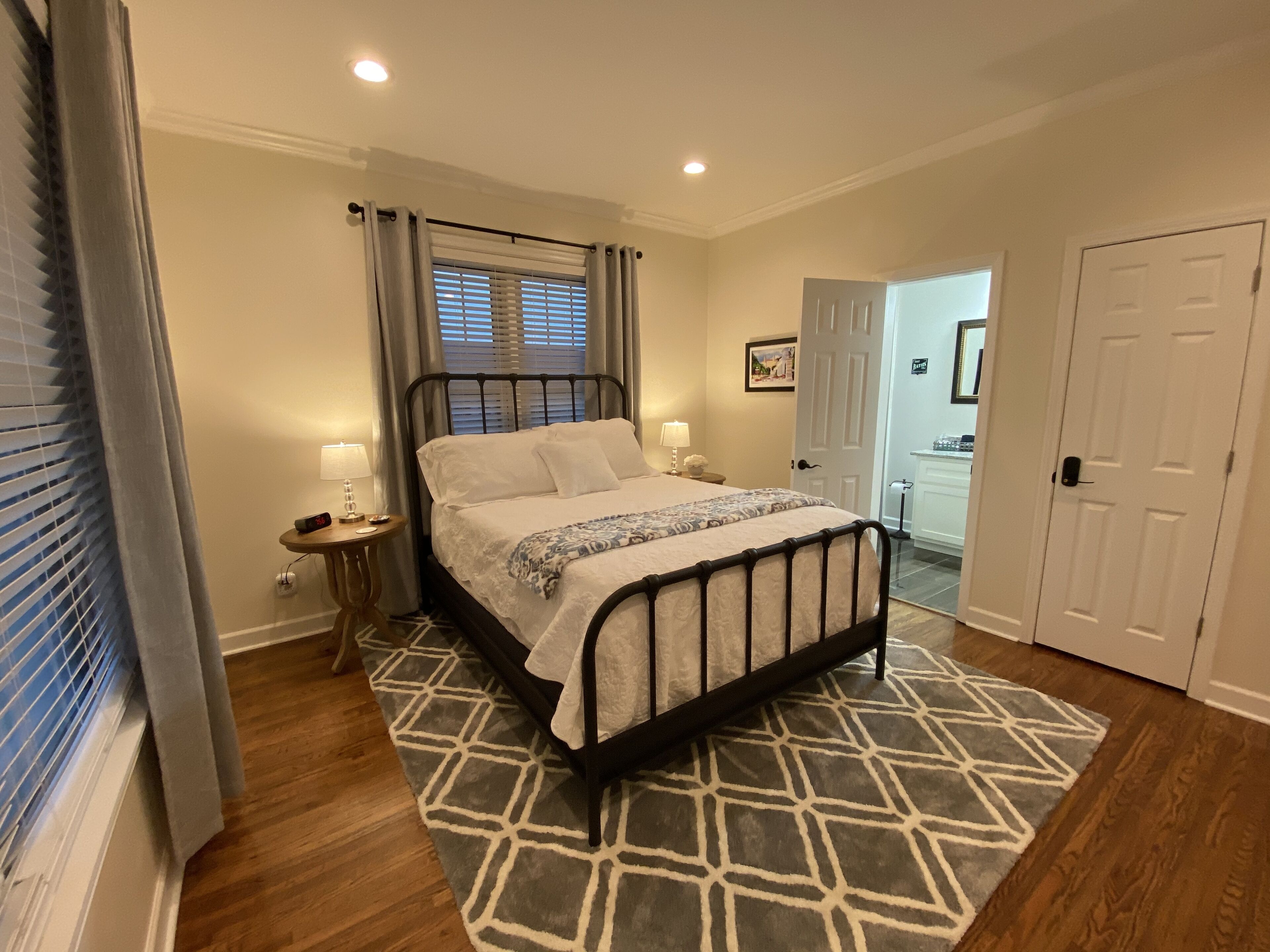 A neatly arranged bedroom with a bed, bedside tables, a patterned rug, and a door leading to another room.