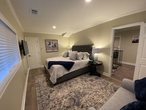 Un dormitorio con una cama grande, una mesita de noche con lámpara, un cuadro en la pared y una puerta que da a otra habitación.