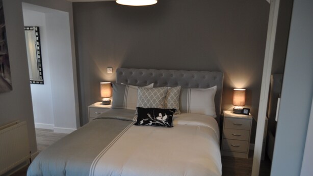 A neatly made bed with a tufted headboard, two bedside tables with lamps, and a nightstand with a clock. - VIP Church Street Apartments (Craigavon)