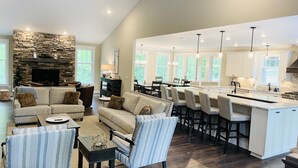 A spacious living room with a stone fireplace, a dining area with high chairs, and a kitchen with a large island and bar stools.