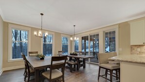 A spacious dining area with a large wooden table and chairs, a chandelier, and large windows offering a view of trees.