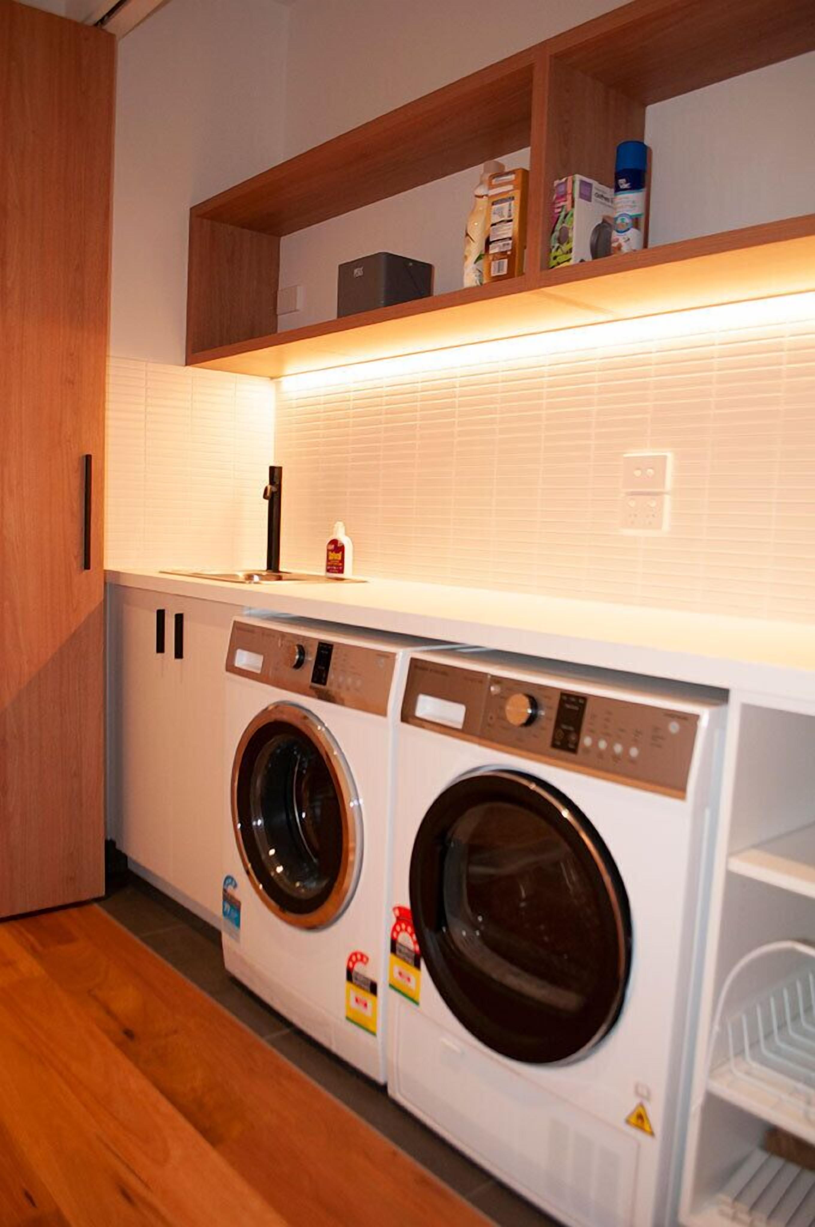 A compact laundry room with two front-loading washing machines, a wooden shelf above, and a tiled backsplash.