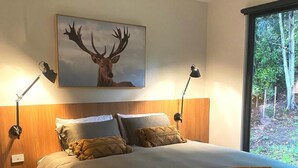 Een moderne slaapkamer met een groot bed, twee bedlampen, een lijst met een herten afbeelding, en een raam met uitzicht op bomen.