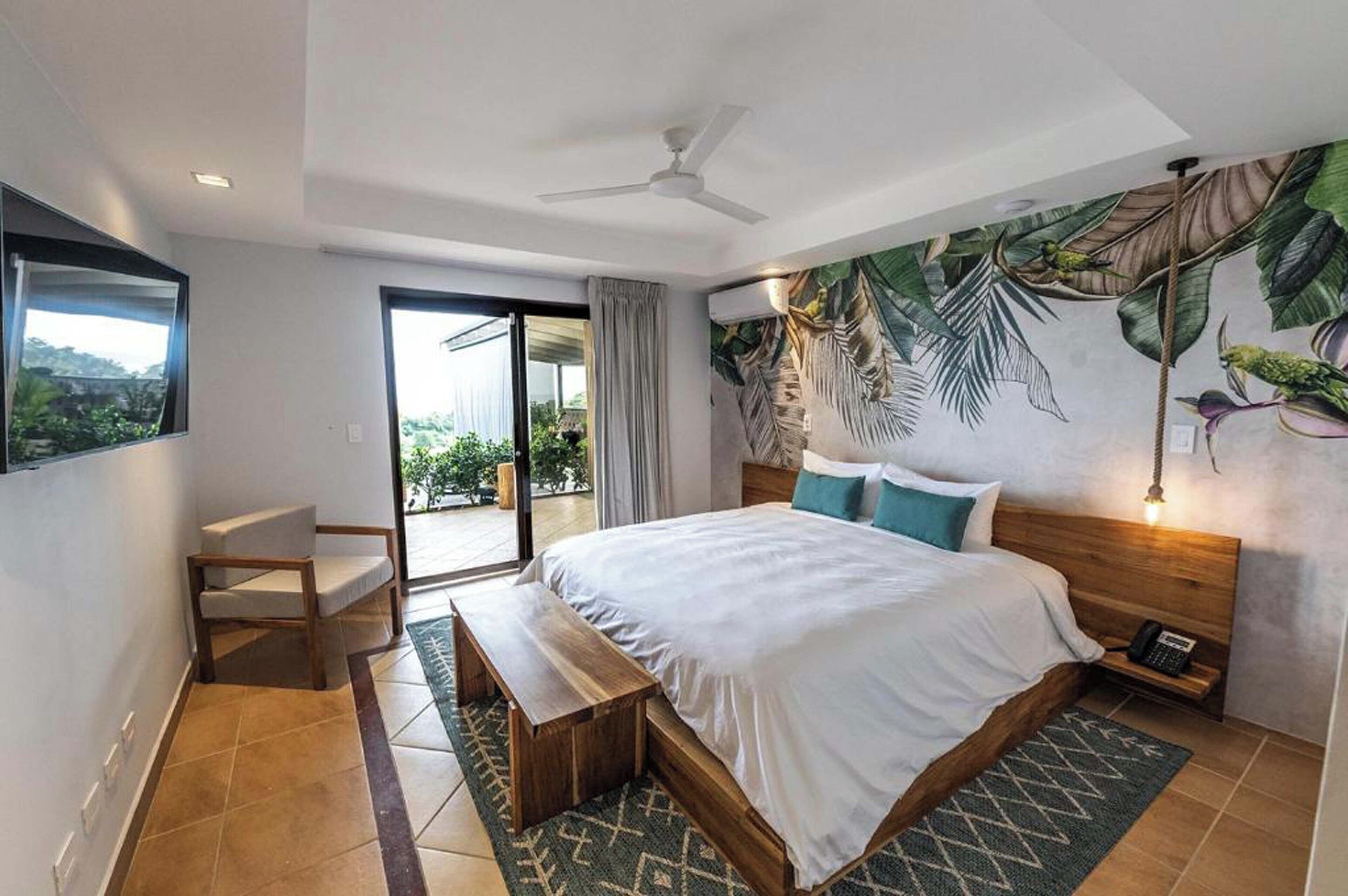 Een moderne hotelkamer met een groot bed, een bank, een plafondventilator, uitzicht door een glazen deur en een tropische muurschildering.