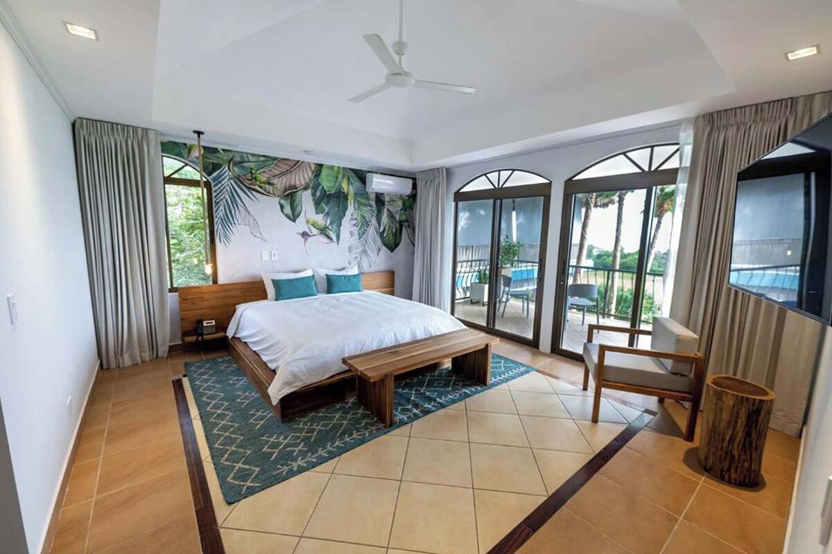 A modern hotel room with a large bed, a wooden bench, a small table, and a view of the pool through the glass doors.