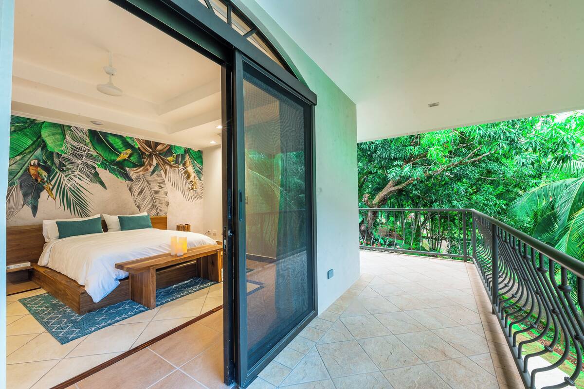A bedroom with a large bed, a bedside table, and a view of lush greenery through a sliding door.