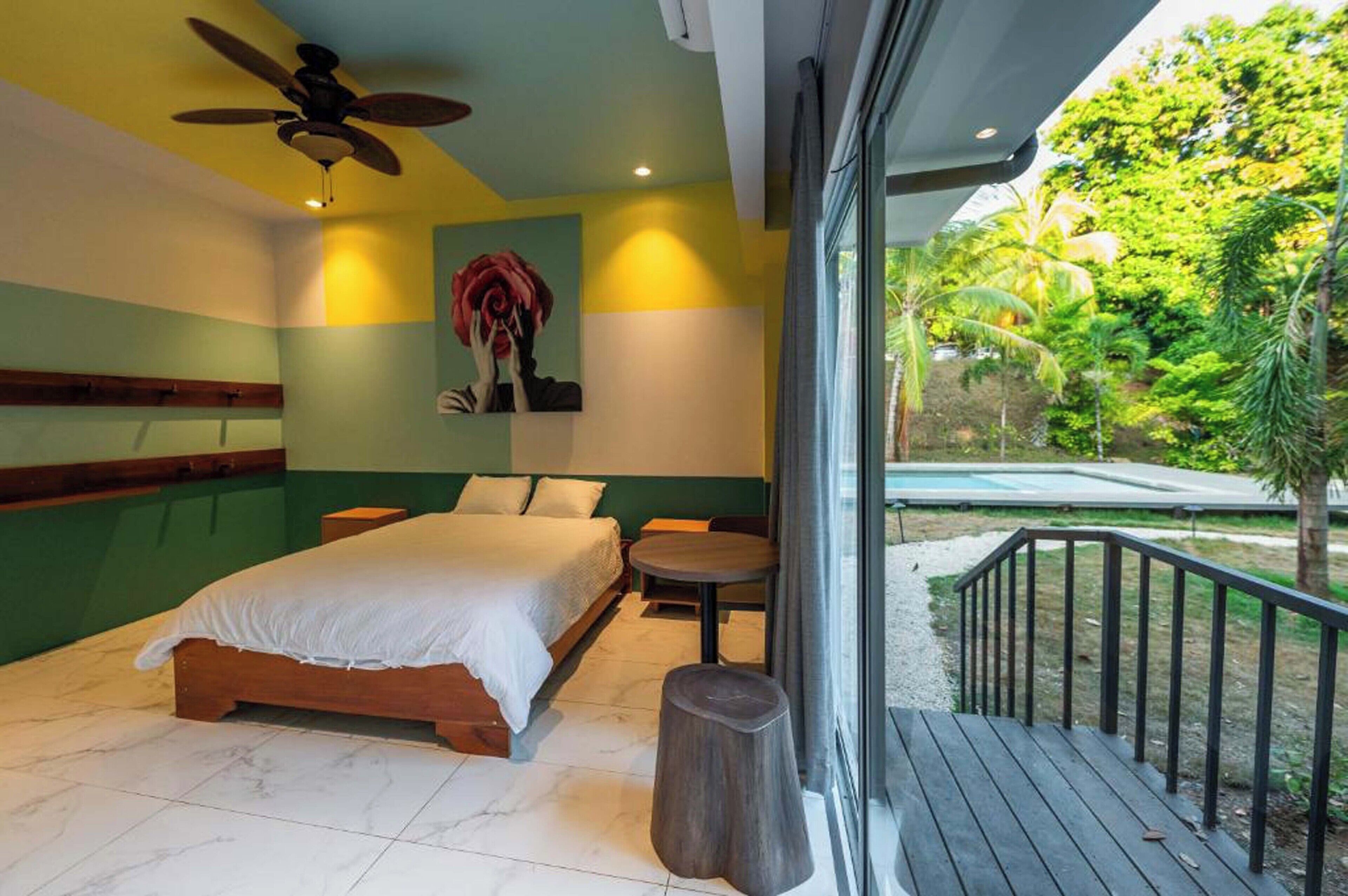 Une chambre avec un lit, un ventilateur de plafond, une petite table et une vue sur une piscine et la verdure.