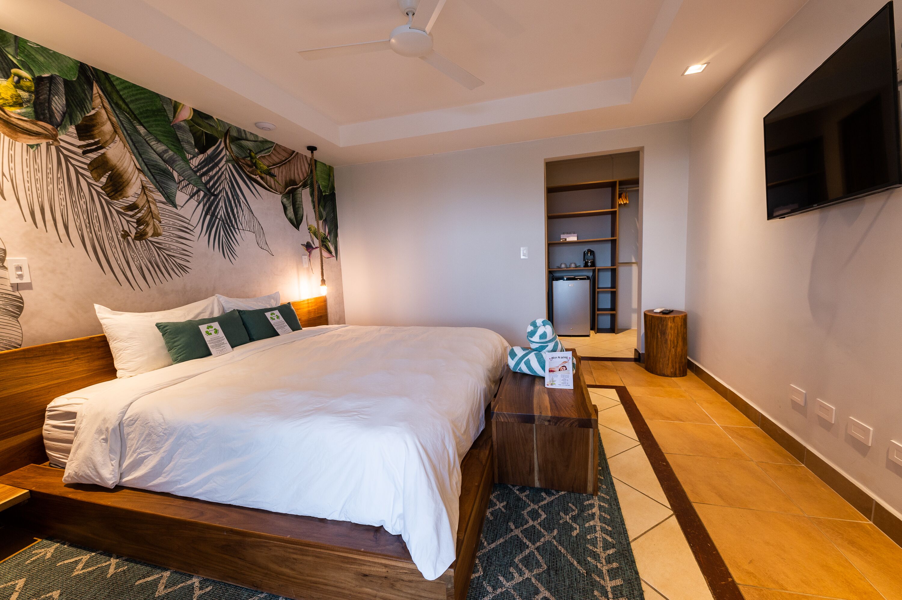 A modern hotel room with a large bed, a flat-screen TV, a ceiling fan, and a tropical wall mural.
