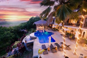 Zona junto a la piscina con sillones de descanso y sombrillas, rodeada de exuberante vegetación y vistas al paisaje.