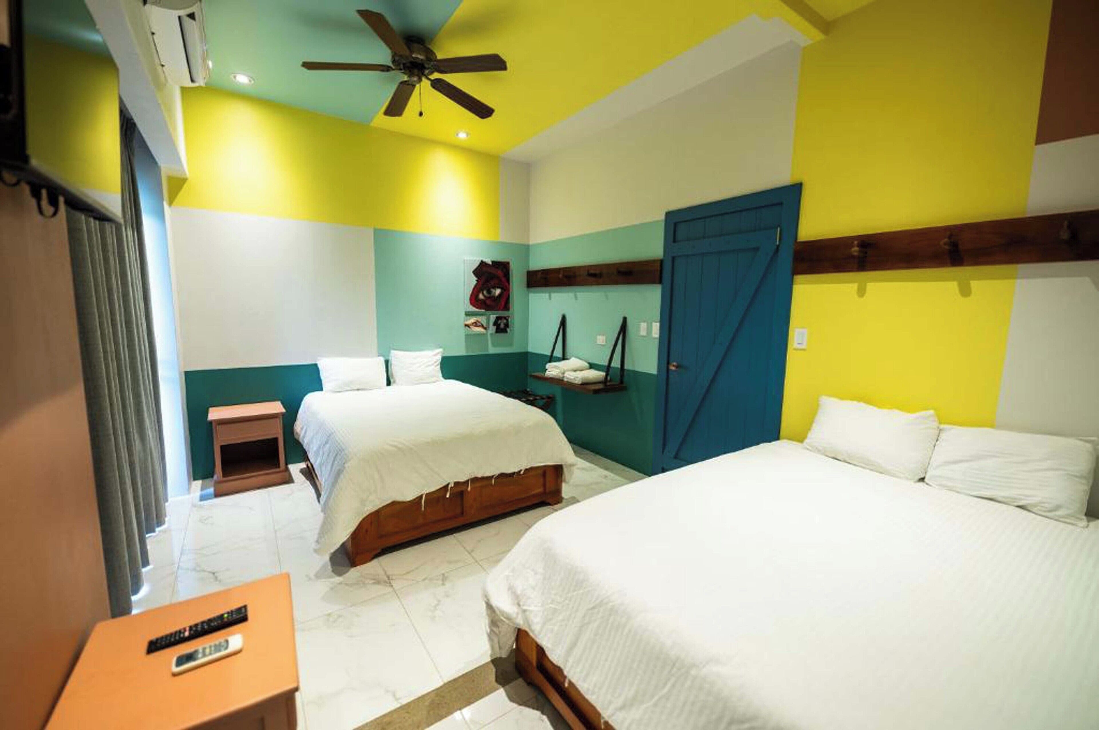 Habitación de hotel con dos camas, un ventilador de techo, una mesita con un control remoto y una puerta azul.